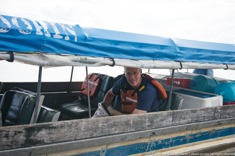 Adult man in a flotation vest sits inside a narrow wooden motorboat with a rolled blue canopy and gear behind him.