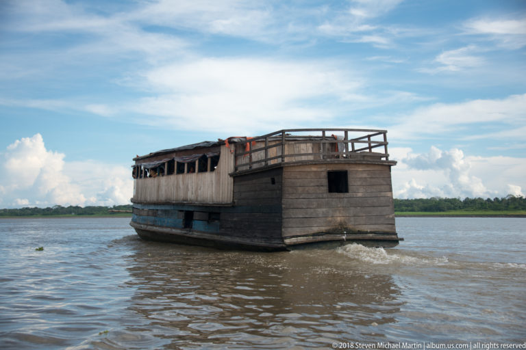 Large weathered two-level wooden riverboat with dark plank hull and open upper railings on a wide river.