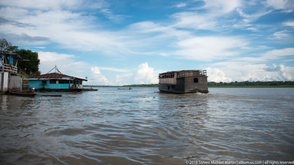 Weathered wooden riverboat offshore from low riverside structures, with a small canoe on the broad channel beneath blue sky.