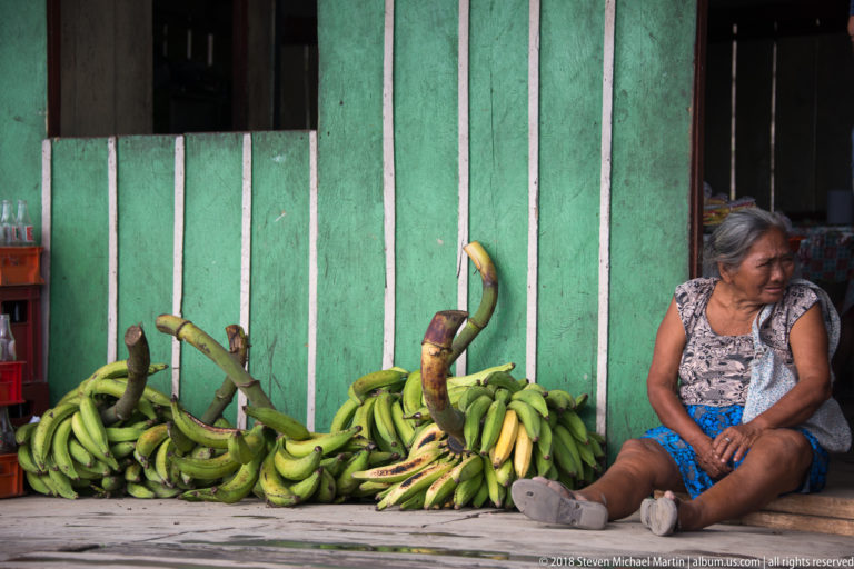 Older woman sits on a wooden platform beside several large bunches of green plantains against a green wall.