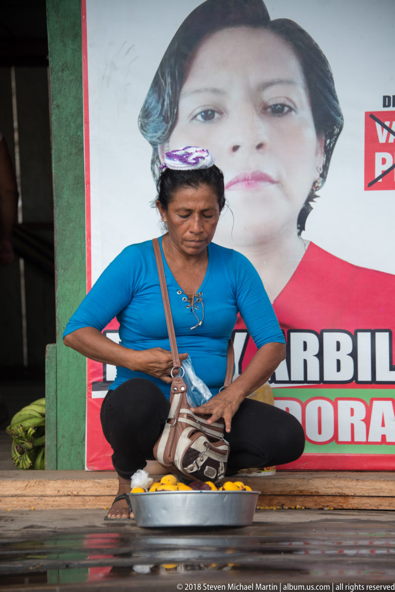 Woman seated beside a metal basin at a riverside storefront, with plantains and a large printed poster behind her.