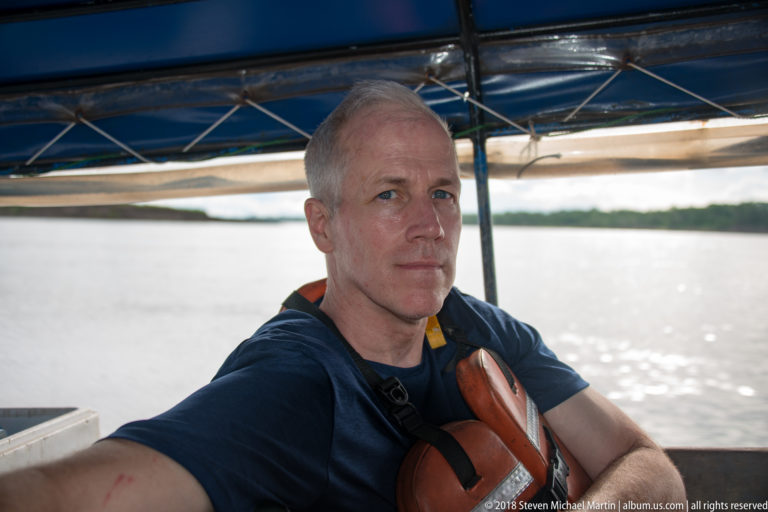 Adult man wearing a flotation vest sits beneath a blue motorboat canopy with the river blurred behind him.