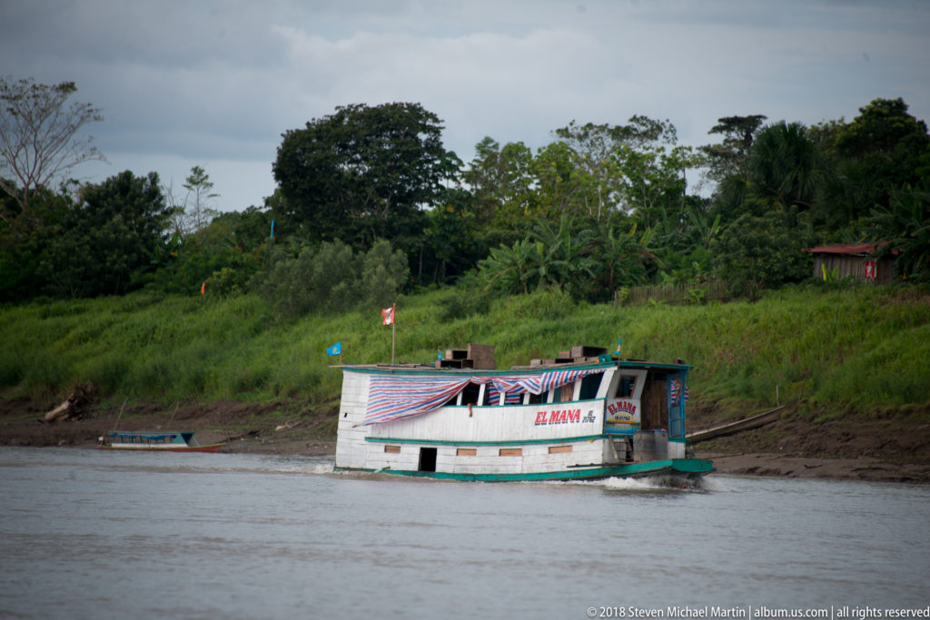 Small white-and-green enclosed riverboat moored against a muddy, vegetated Amazon riverbank.
