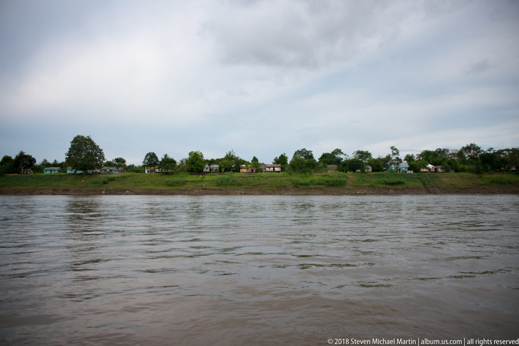 Wide river in the foreground with a small line of houses on a low green bank beneath a cloudy sky.
