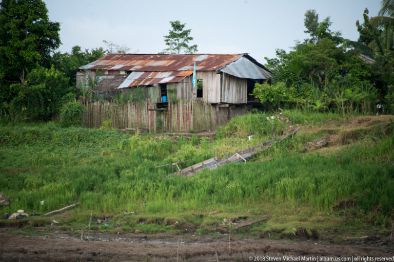 Weathered wooden riverside house with corrugated-metal roof, picket fence, and a narrow canoe on the grassy bank below.