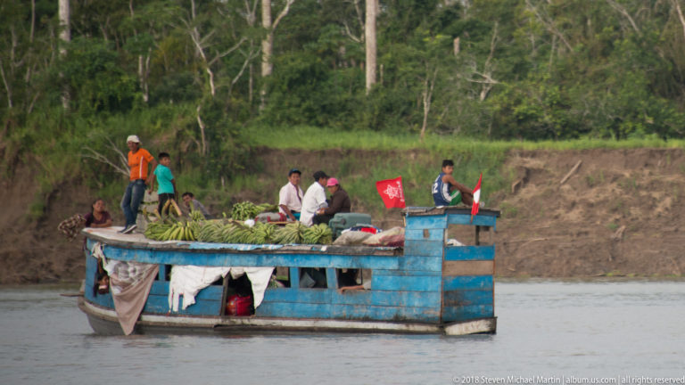 Weathered blue riverboat carrying passengers, green plantain bunches, and bundled goods along a muddy Amazon channel.
