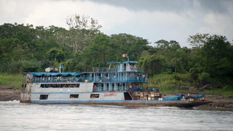 Multi-deck riverboat moored beside a low forested bank on a broad brown-water channel in the Peruvian Amazon.