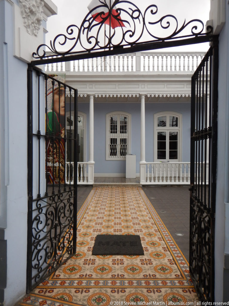 Open wrought-iron gate framing a patterned tile courtyard and pale blue veranda facade.
