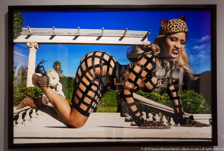 Woman in a black lattice bodysuit with leopard-pattern accessories poses on all fours on a sunlit classical terrace.