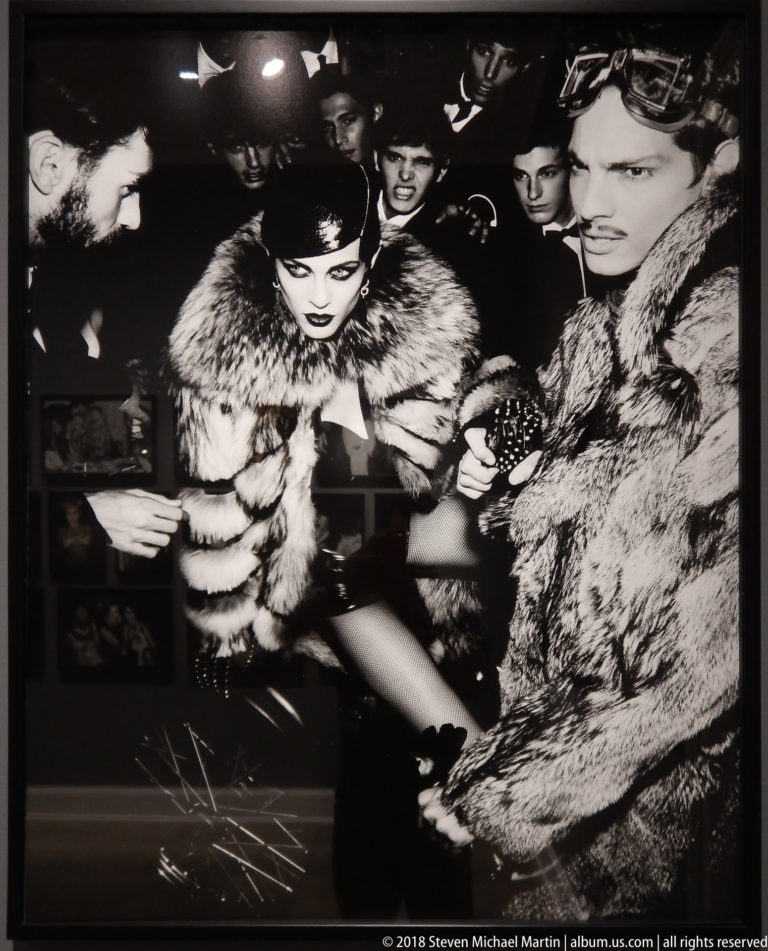 Crowded black-and-white party scene with a central woman leaning forward among figures wearing dark clothing and fur.