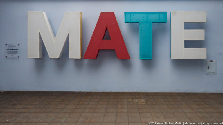 Large dimensional letters spelling MATE mounted on a pale blue wall above a tiled floor.