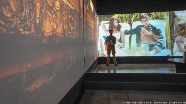 Visitor silhouetted on a platform before large color fashion projections wrapping a dark gallery.