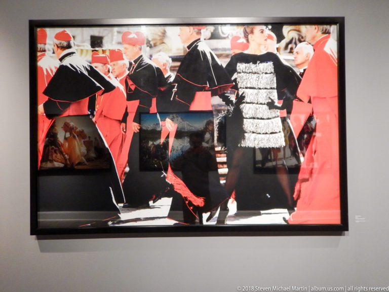 Woman in a textured black-and-white dress stands among men in red-and-black ceremonial clerical clothing.