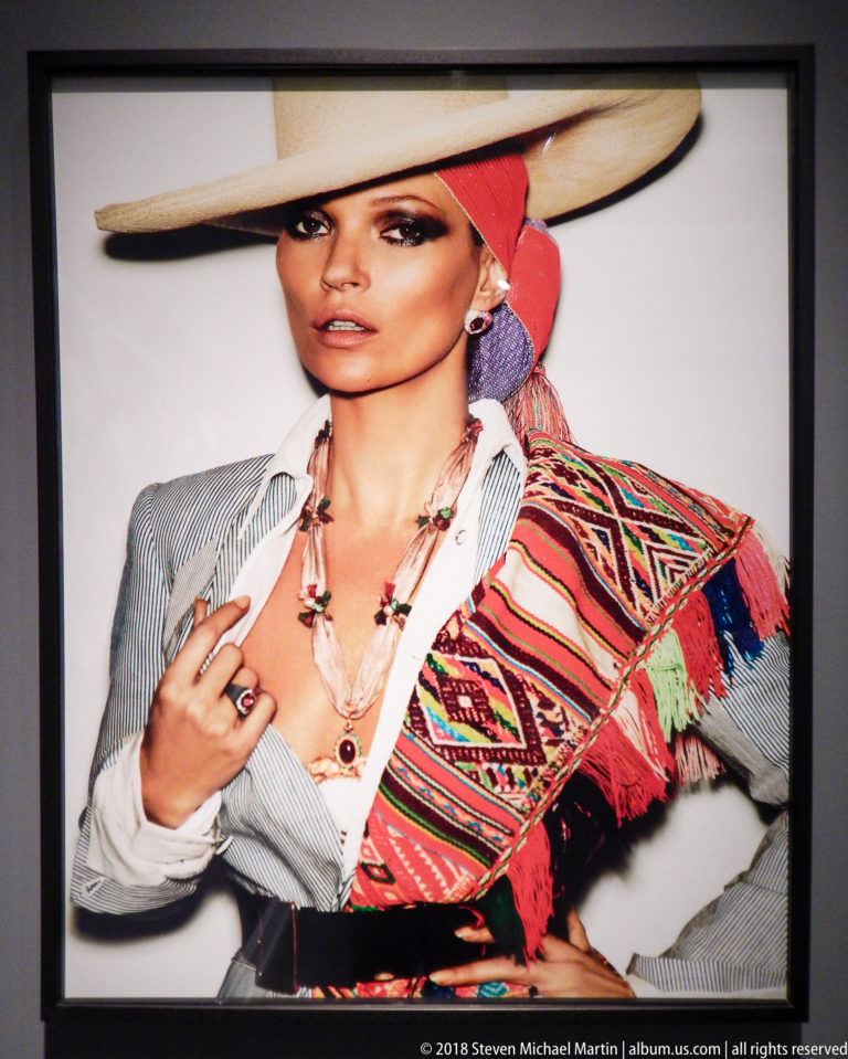 Woman in a broad hat and striped shirt wears layered jewelry and a bright woven textile over one shoulder.