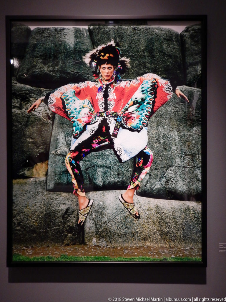 Woman in a vivid patterned outfit poses with bent knees and outstretched arms against a rough stone wall.