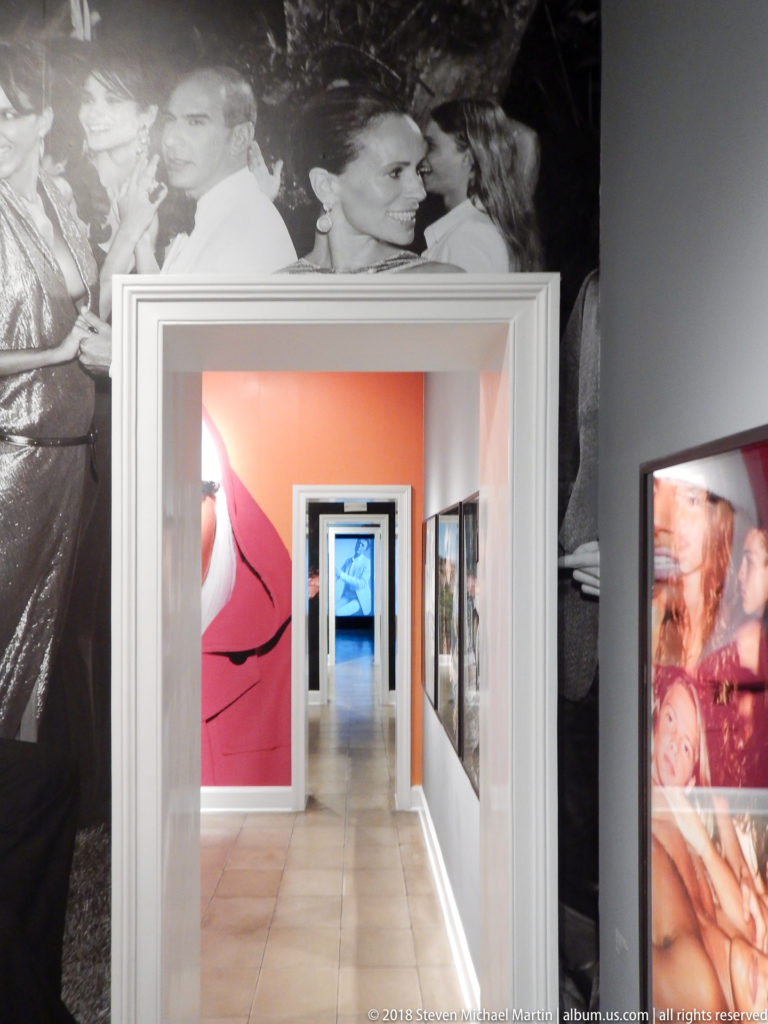 White doorway framing a long gallery corridor with coral walls and fashion photographs; large works surround the entrance.