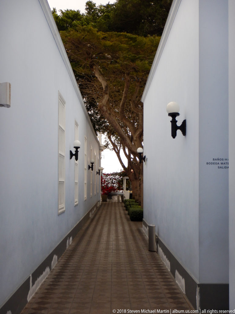 Narrow walkway between pale blue museum buildings with black wall lamps, white windows, and trees ahead.
