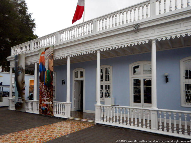 Blue-and-white courtyard facade with veranda, white balustrade, patterned tile paving, exhibition banners, and a Peruvian flag.