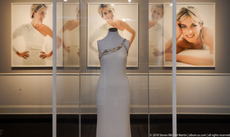 Pale floor-length evening dress in a glass case with large portraits of Diana, Princess of Wales, behind it.