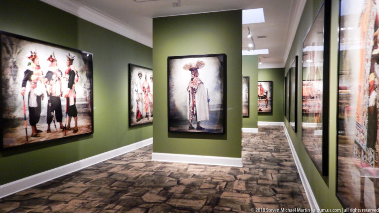 Green museum gallery lined with large portraits of traditional Peruvian dress, seen through a central partition and reflection.