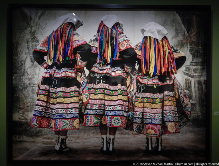 Three women from behind wearing embroidered traditional dresses with broad skirts and long multicolored ribboned headdresses.