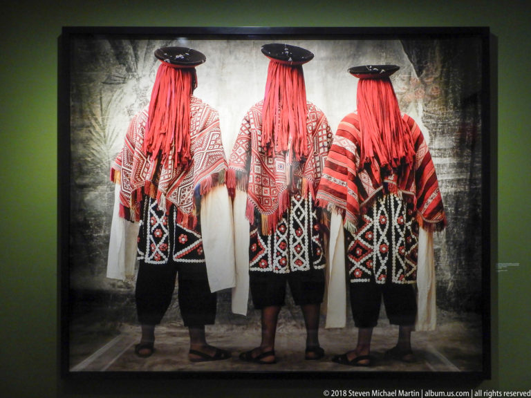 Three figures seen from behind wearing broad hats and coordinated red, black, and white traditional garments.