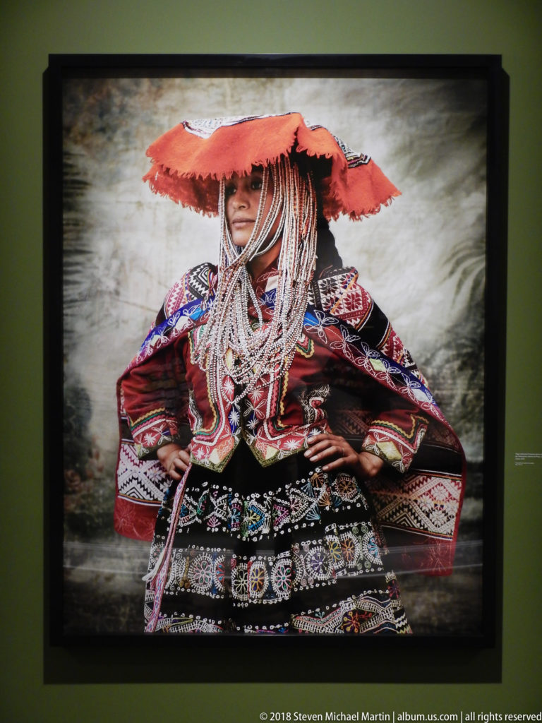 Seated figure in richly embroidered Peruvian dress with layered necklaces, broad red headpiece, and hands at the waist.