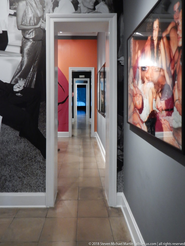 Gallery corridor with tiled floor, nested doorways, coral walls, and large fashion photographs along both sides.