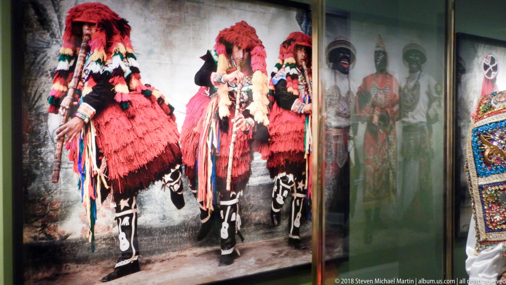 Three figures in red feathered headpieces and black-and-white traditional costumes pose frontally in a large portrait.