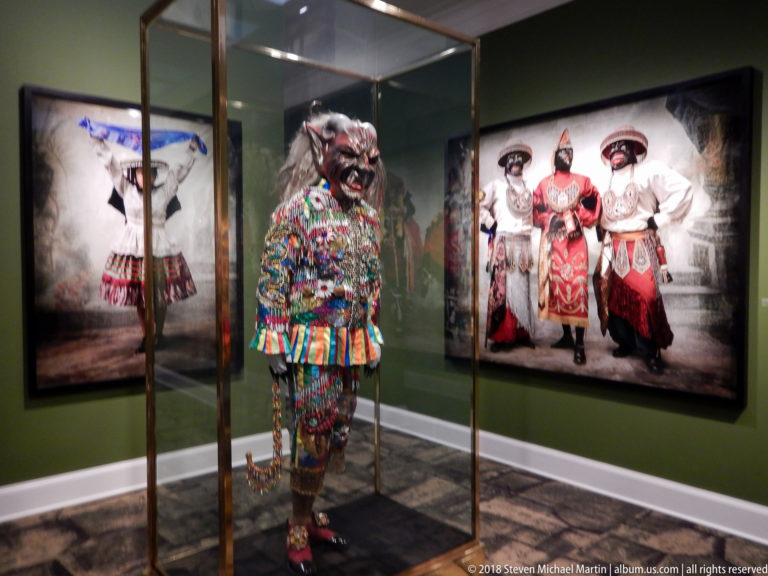 Elaborately beaded and embroidered Peruvian festival costume displayed in a glass case between large costume portraits.