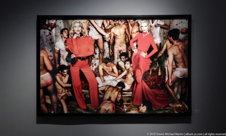 Two women in vivid red outfits stand amid a crowded staged party scene in a large framed color photograph.