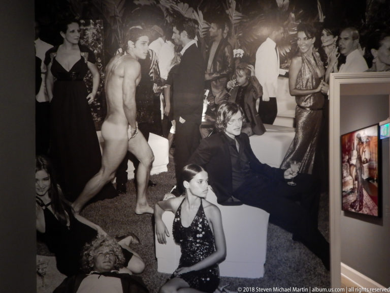 Large black-and-white fashion photograph of a crowded social scene installed beside a doorway and neighboring color work.