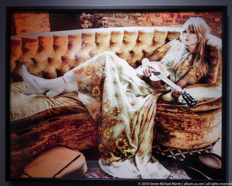 Woman in a pale floral outfit reclines on a tufted sofa holding a dark firearm-like prop across her lap.