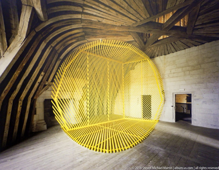 Large yellow radiating elliptical line sculpture filling a rustic timber-roofed interior.