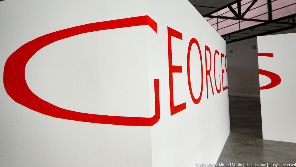 Oversized red word GEORGES wrapping around the corner of white gallery partition walls.