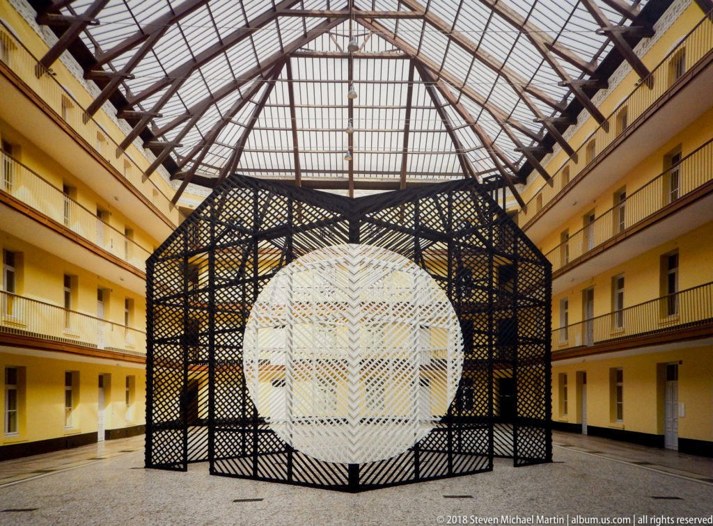 Black lattice enclosure containing a white striped spherical form in a yellow multi-level atrium.