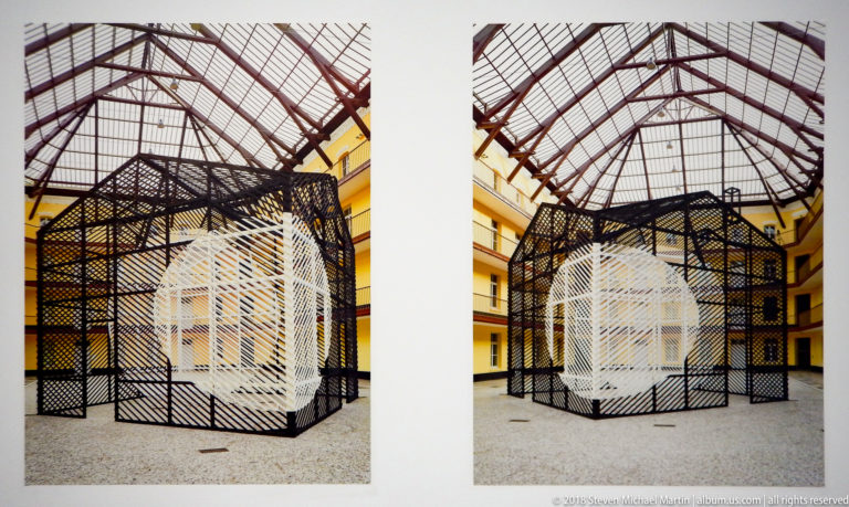 Two views of a black lattice enclosure surrounding a white striped geometric inner form beneath a glass roof.