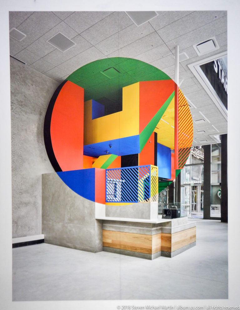 Large oval geometric installation in red, green, yellow, blue, and black spanning a bright museum interior.