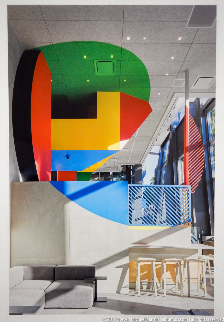 Side view of multicolored geometric oval installation above seating and tables in a bright interior.
