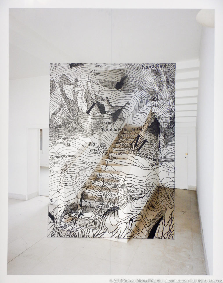 White gallery with a cube and wall panel covered in dense black cartographic linework and place names.