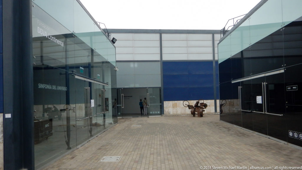 Paved passage between modern glass-fronted museum buildings, with blue wall panels and a small sculpture at the far end.