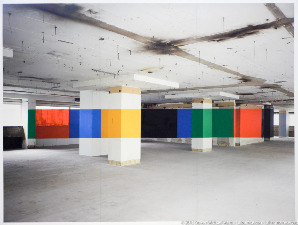 Wide unfinished room with columns and walls painted in long red, blue, yellow, black, green, and white bands.