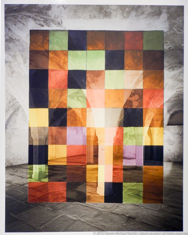 Grid of translucent multicolored rectangular panels filtering a view into a rough interior.