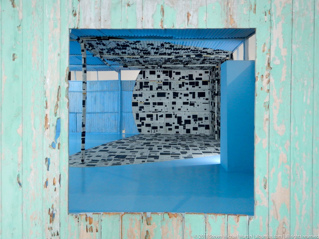Peeling turquoise wall opening framing a black-and-white installation and bright blue floor beyond.