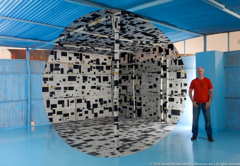 Black-and-white patterned cube installation in a blue room, with a standing visitor at right.