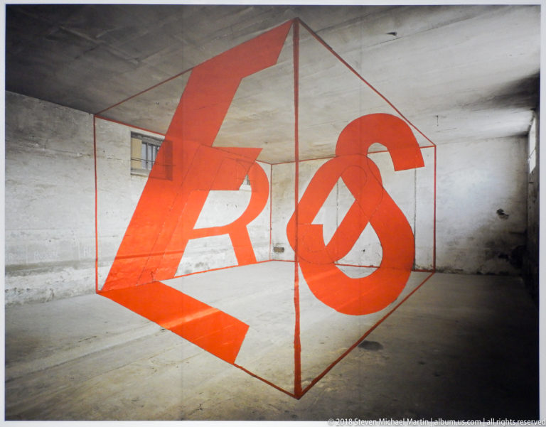 Red perspective-based letter forms aligned inside a wireframe cube in a worn white room.
