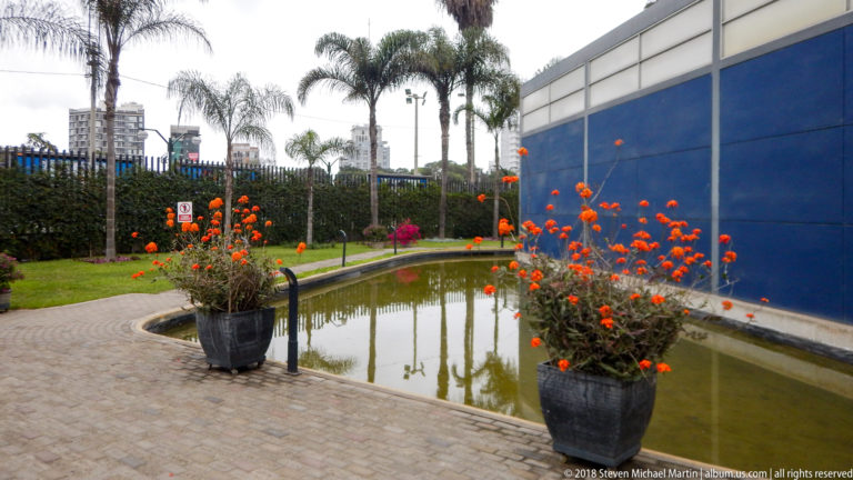 Courtyard reflecting pool bordered by orange flowering plants, palms, paving, and a tall blue museum wall.