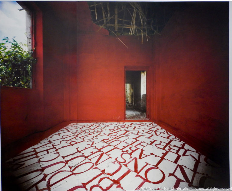 Red room with a central doorway and oversized fragmented red letterforms across a white floor.