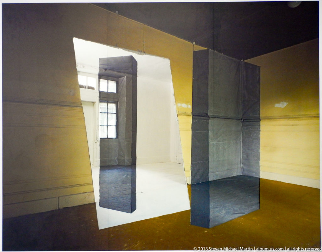 Dark gray and yellow geometric installation with suspended rectangular forms around a bright central opening.
