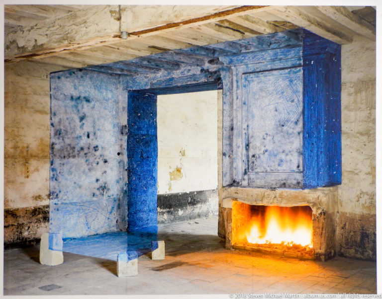 Rough blue-painted doorway-like installation beside a low rectangular open flame in a weathered interior.